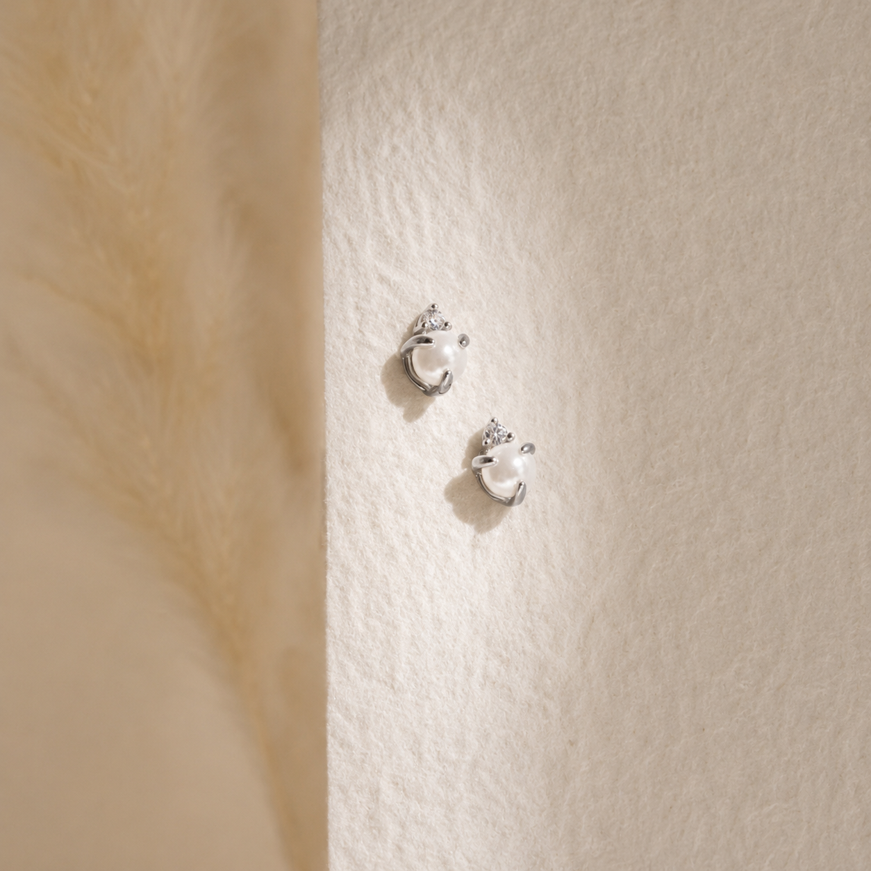 A pair of silver crescent moon stud earrings with small clear crystal accents, displayed on a textured cream wall next to a soft beige furry surface.